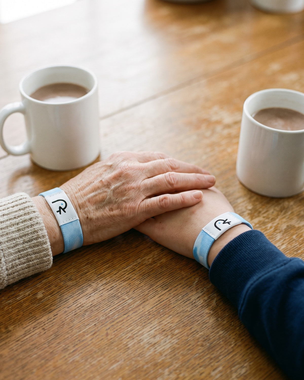 A grandmother’s hand resting on her granddaughter’s hand between two mugs of cocoa, both wearing Faith Over Fear bands