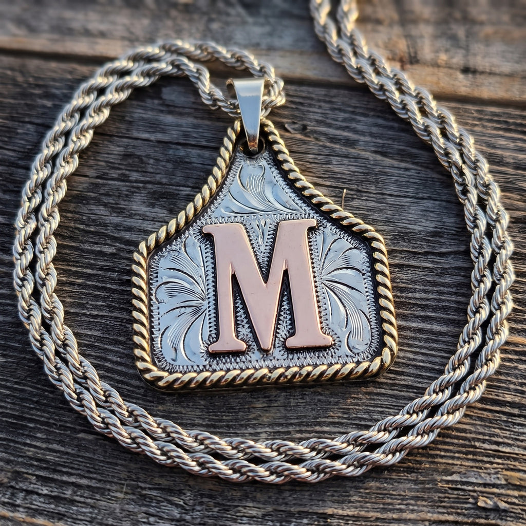 Cow Tag Initial Necklace & Keychain – engraved silver cow tag with copper letter M on a rope chain, on weathered barn wood