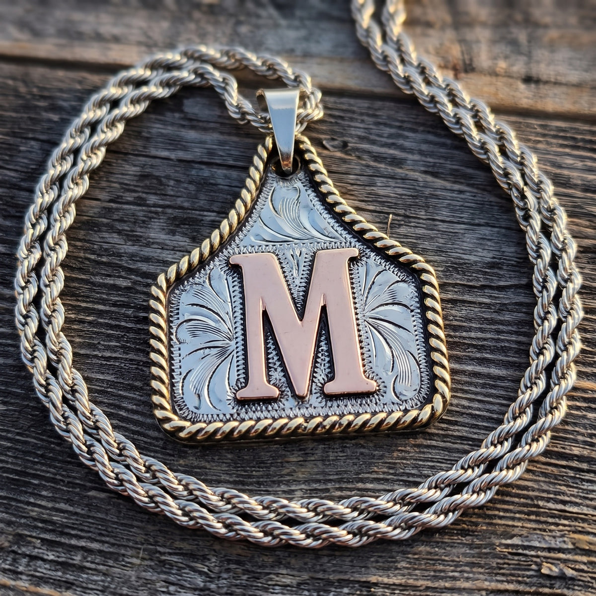 Cow Tag Initial Necklace & Keychain – engraved silver cow tag with copper letter M on a rope chain, on weathered barn wood
